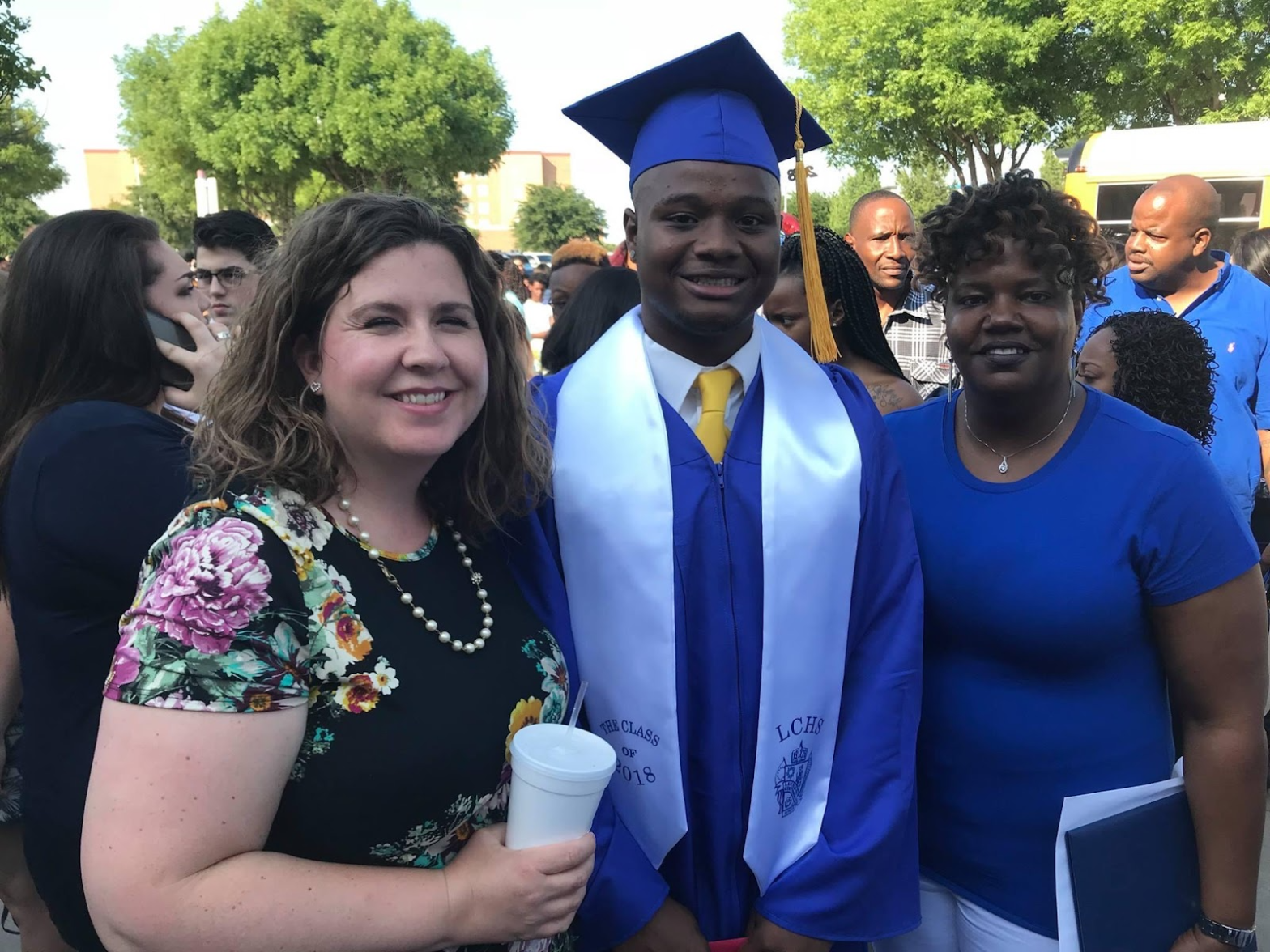 A white woman in a floral dress holding a white cup. Her arm is around a young, black male and his mom, a black woman. The male and mom are both dressed in blue. The male has his graduation cap and gown on. They are celebrating after he graduated.